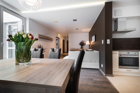 A kitchen table with flowers in the center, and part of the kitchen and living room in the distance.