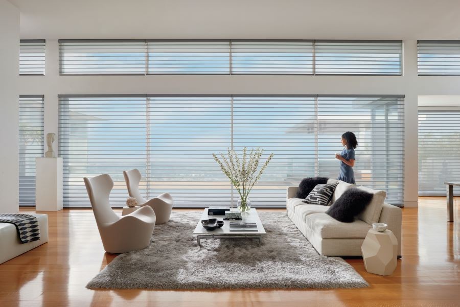 A woman standing in a living room looking out picture windows covered by Hunter Douglas sheer shades.